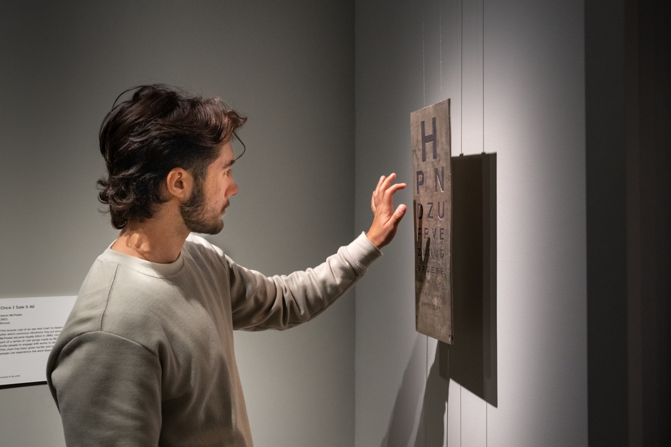 Photograph of an individual exploring an exhibition. They are stood facing a gallery wall. In front of the wall a rectangular bronze eye test chart with the standard arrangement of letters is hanging, secured by 4 wires. The individual has their hand raised as if they are about to tap the bronze chart with their finger.