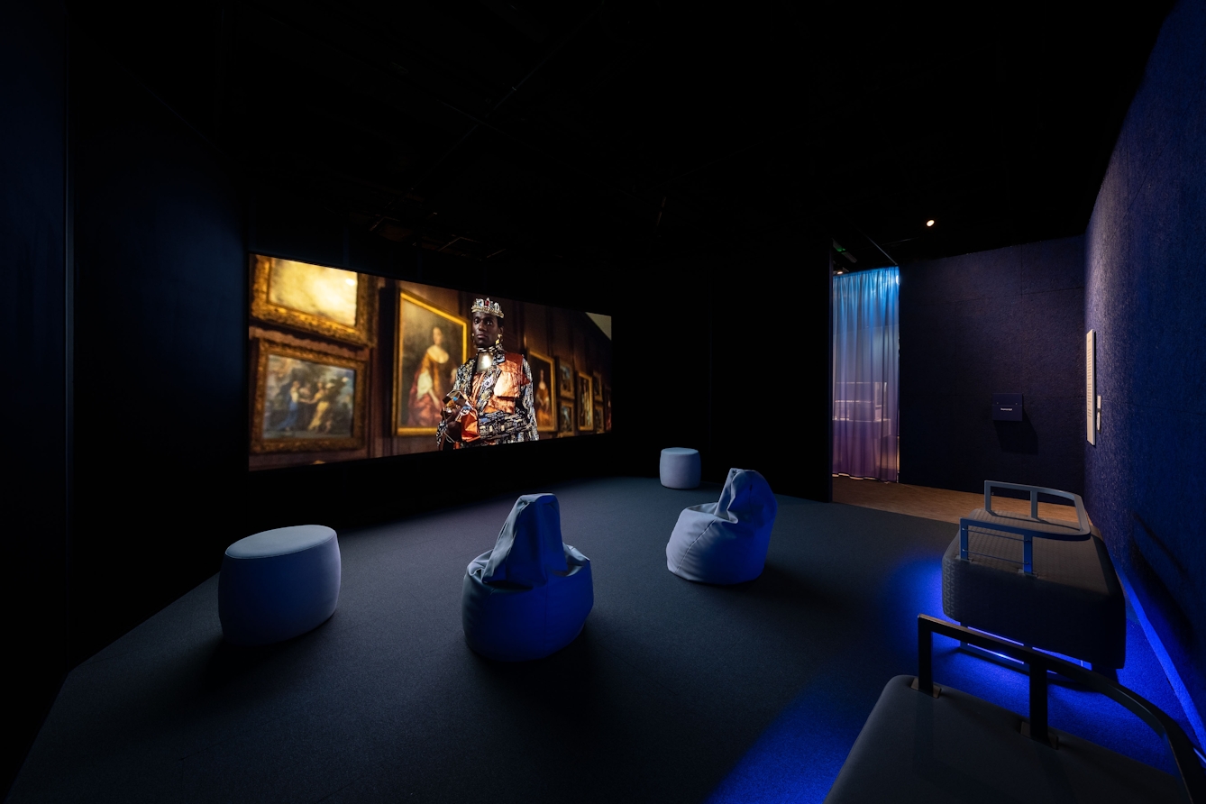 A scene from a video plays on a wall in a darkened room with different seats and beanbags scattered around the room. The scene being broadcast is of a black man in an oppulent constume wearing a crown. Behind him is a wall of framed historical European paintings.