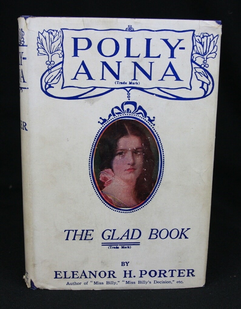 Photograph of a book cover of 'Pollyanna'. Text on the cover reads 'Pollyanna, The Glad Book by Eleanor H. Porter'. 

The book cover is white with blue writing. In the middle of the cover is an inset drawing of a young woman in a frame. 