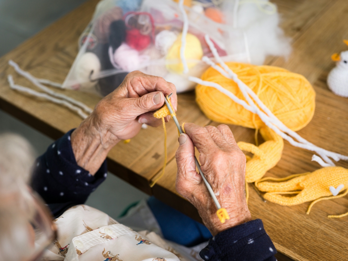 A photograph of the hands of an elderly woman knitting. They are holding two grey knitting needles and are making stitches with yellow wool. In the background is a ball of yellow wool and a bag containing small balls of wool in various colours.