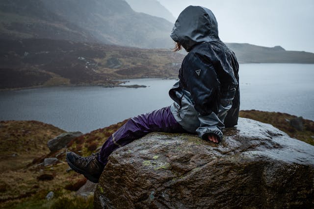Photograph of a young child sitting on a rock wearing a wet raincoat, trousers and walking boots. Their back is to the camera and they are facing a lake and several mountains. 