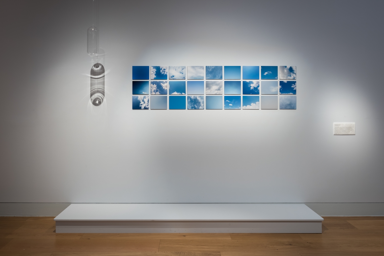 Colour photograph of an exhibit in a museum. There are 27 small images of the sky, most of them blue and many with white clouds. The images are all positioned close together on a white wall. To the bottom right of the images is a small white textbook . 