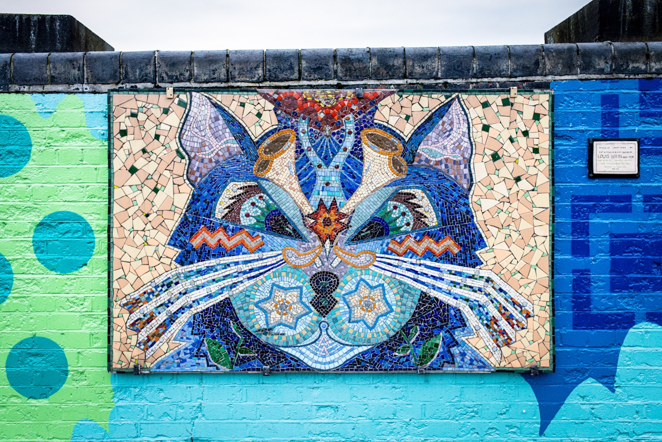 Photograph showing a brightly coloured abstract ceramic mosaic of a cat, in the style of Louis Wain. The mosaic is hung on a low brick wall which is also brightly coloured. Above the wall, a thin slither of sky can be seen. To the right side is an information plaque. 