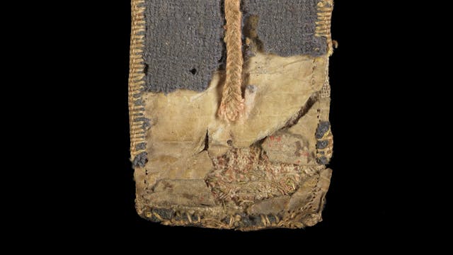 Photograph of a 15th century Medieval folding almanac. The embroidered binding is worn and there is an exposed layer of parchment below, with faint text lettering on it. 