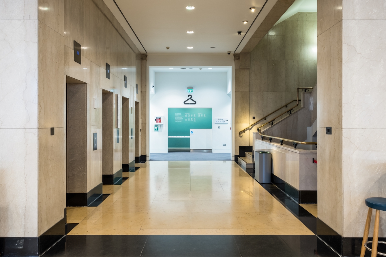 A photograph of the first floor facing four lifts on the left, a staircase going downwards and upwards on the right, and a cloakroom directly opposite  had you come straight in with from Euston Road with the entrance behind you.