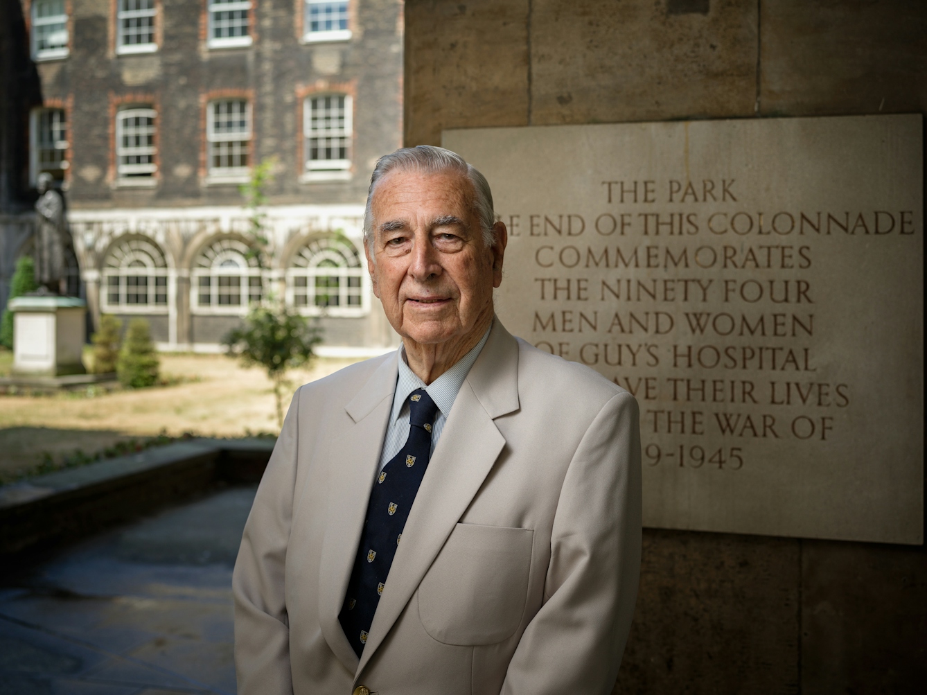 Photographic portrait of a man standing in the colonnades at Guys Hospital, London. Behind him is a stone plaque commemorating the hospital staff that lost their lives during the Second World War.