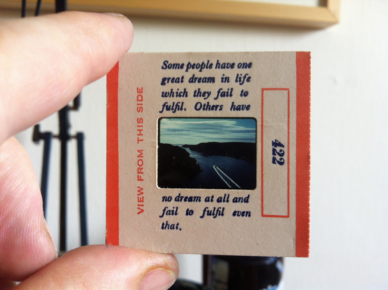 A person holds an old-fashioned slide showing an image of a boat travelling down a river. The slide has been printed with an extract from The Book of Disquiet.