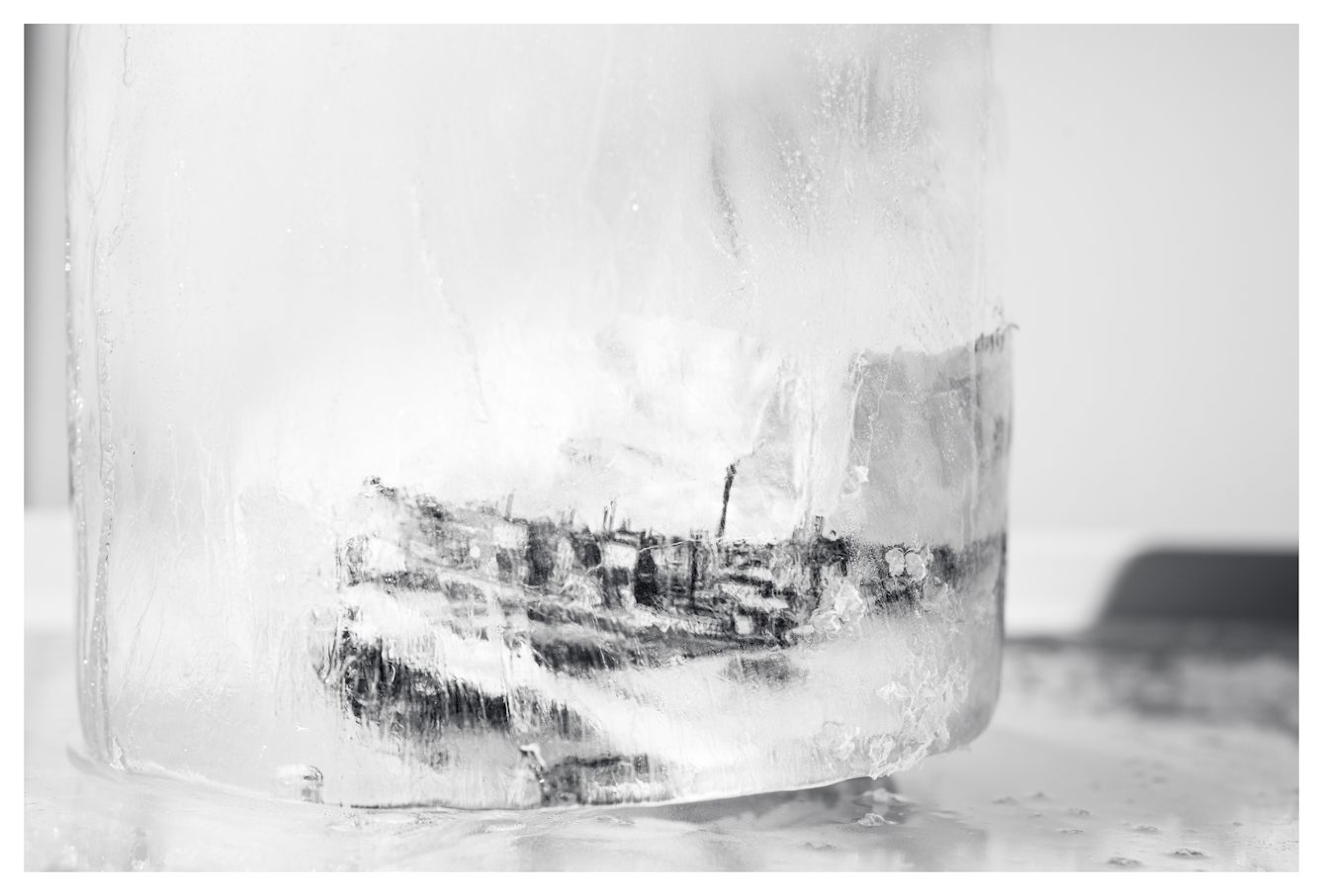 Monotone photograph showing a cylindrical core of frozen water. Encased within the ice is an illustration of an industrial town scene from the 1800s that can just be seen through the distortions of the ice wall and opaque frosting. The ice core is standing vertically on a glass fridge freezer shelf.