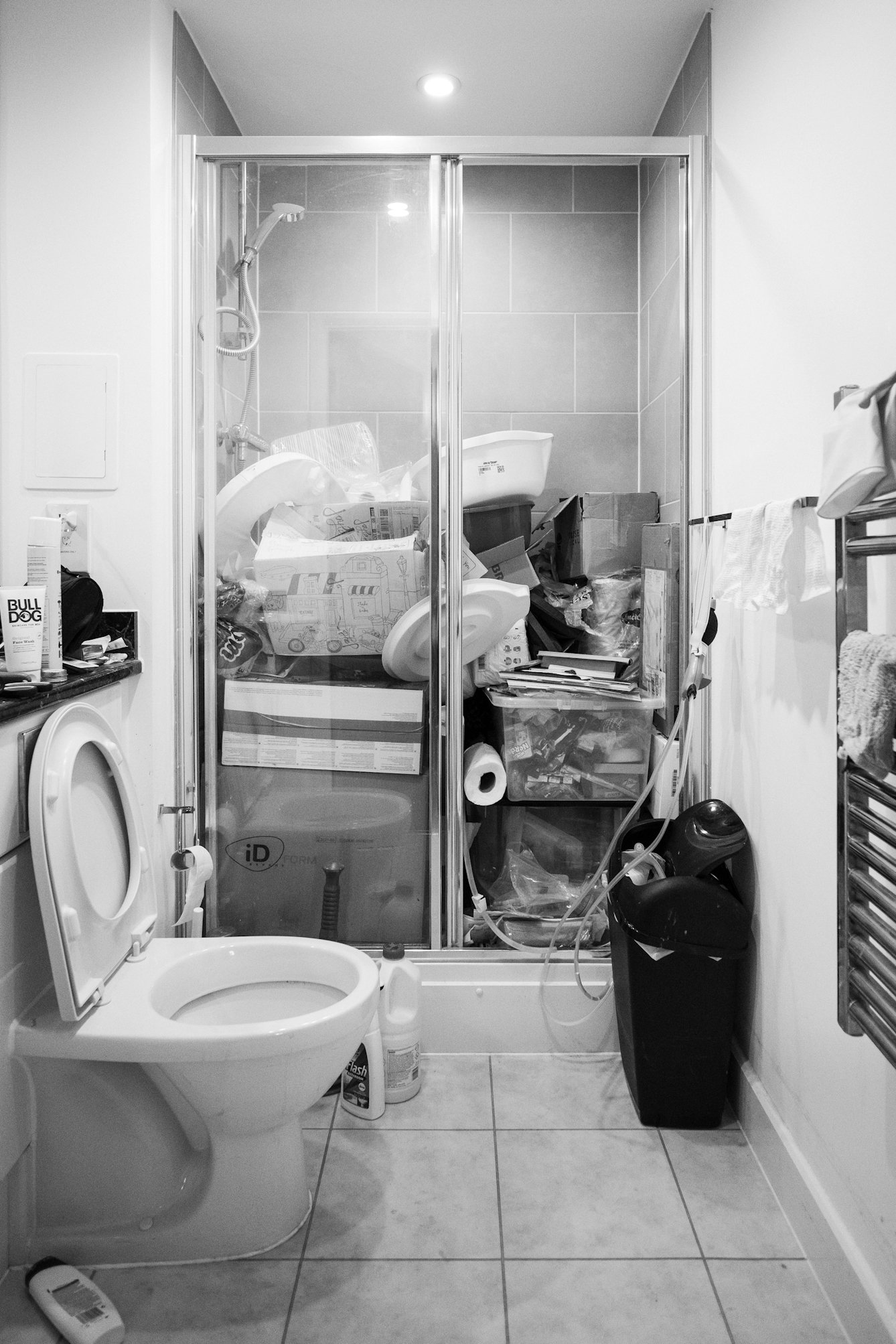 Black and white photograph of a bathroom shower cubicle which is full of objects and being used as a storage area rather than a shower.