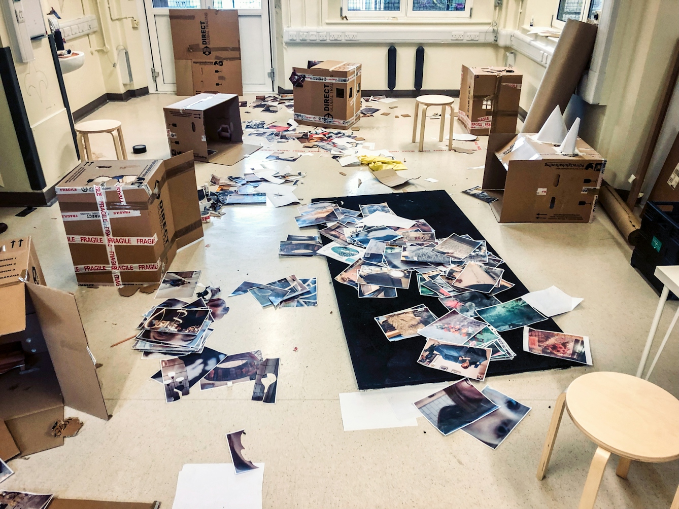 A photograph showing a room with magnolia painted walls, white PVC double doors and windows, and a lino floor on which a multitude of colour photocopies have been cut up and are strewn messily, many of which are on a black rectangular mat in the middle of the room.  Around the edge of the room there are six large open cardboard boxes on their sides, some with fragile stickers. 