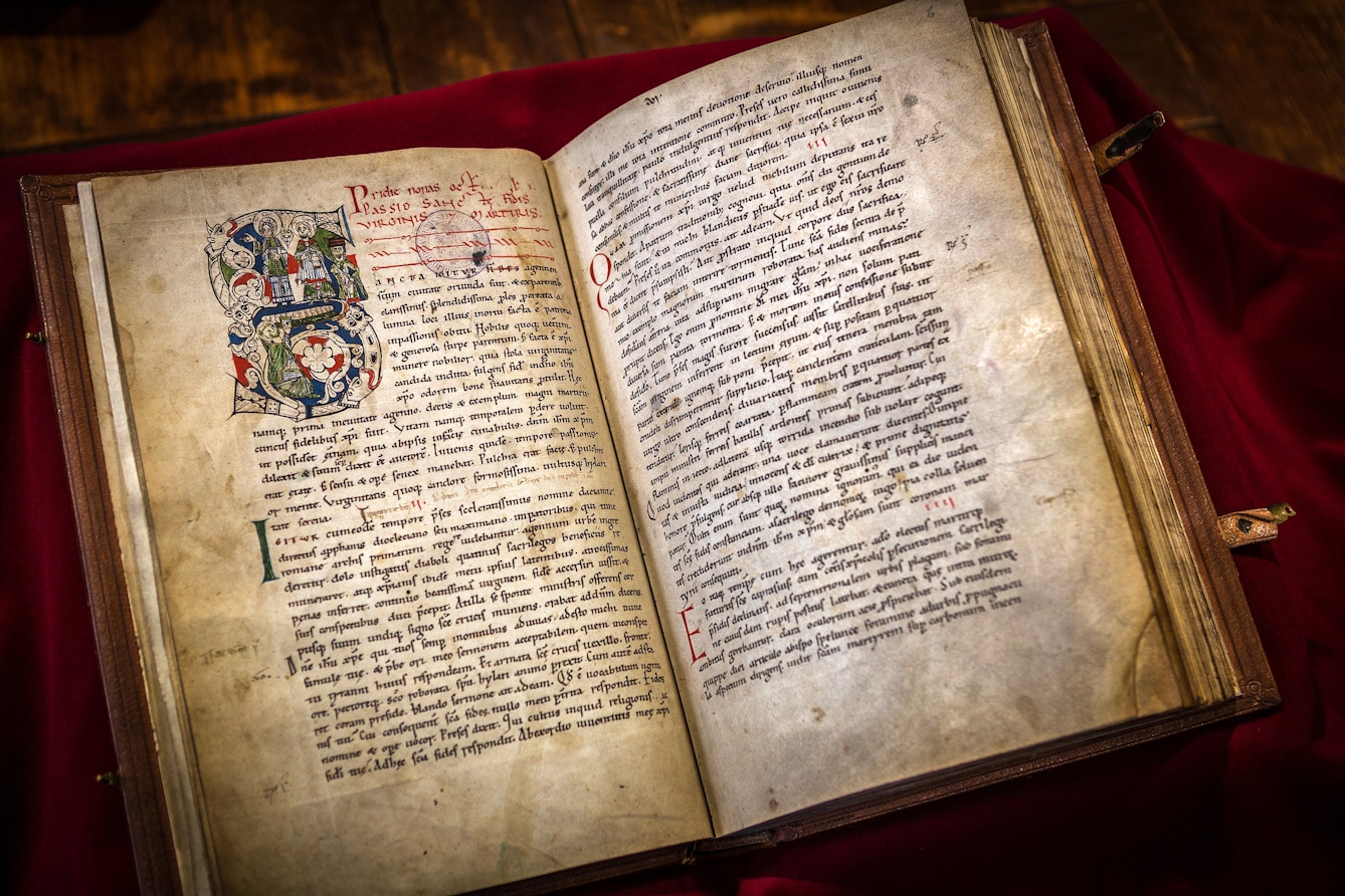 'The Miracles of Sainte Foy' by Bernard of Angers, c. 1013–1020. A medieval manuscript, bound and open showing two pages of text. The left page has a very ornate illuminated lettter S which includes figures of saints, mythical animals and flora.