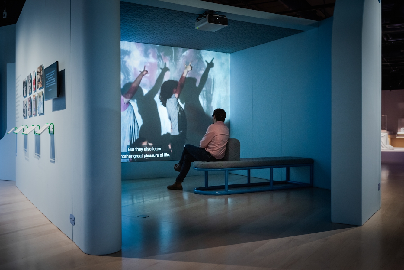Photograph of a man sitting on a bench in an exhibition space watching a projected film. The film still shows 2 children with their hands in the air casting a shadow on the wall behind. The subtitle on the film reads, 'But they also learn another great pleasure of life.'.