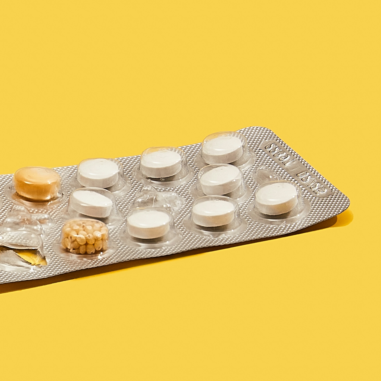 Photograph of a foil medicine pill blister pack sitting on a bright yellow background. The blisters on the right side of the pack contain white tabloid pills. The blisters on the left of the pack contain a single kernel of corn or a collection of small grains of maize.
