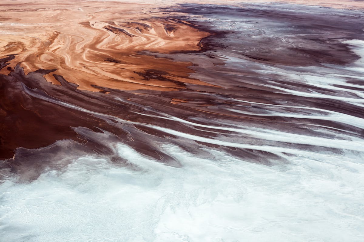 Aerial colour photograph showing the mineral formations at the edges of the salt flats along the Rio Grande delta in Bolivia. The image is abstract in nature making it difficult to get a sense of scale. The overall tones are a brown, red, rust colour with expanses of white. The brown starts in the top left corner swirling in patterns of differing hues across towards the bottom right corner of the image. Half way across this diagonal, the brown begins to mix with the white, merging with long swirling strands, until eventually arriving at the bottom of the image where they turn completely white with a blue green tint and grainy texture.