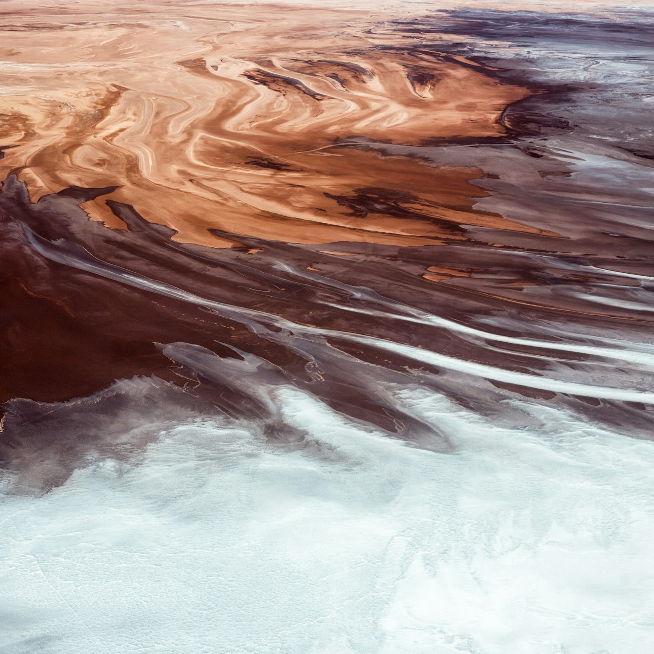 Aerial colour photograph showing the mineral formations at the edges of the salt flats along the Rio Grande delta in Bolivia. The image is abstract in nature making it difficult to get a sense of scale. The overall tones are a brown, red, rust colour with expanses of white. The brown starts in the top left corner swirling in patterns of differing hues across towards the bottom right corner of the image. Half way across this diagonal, the brown begins to mix with the white, merging with long swirling strands, until eventually arriving at the bottom of the image where they turn completely white with a blue green tint and grainy texture.