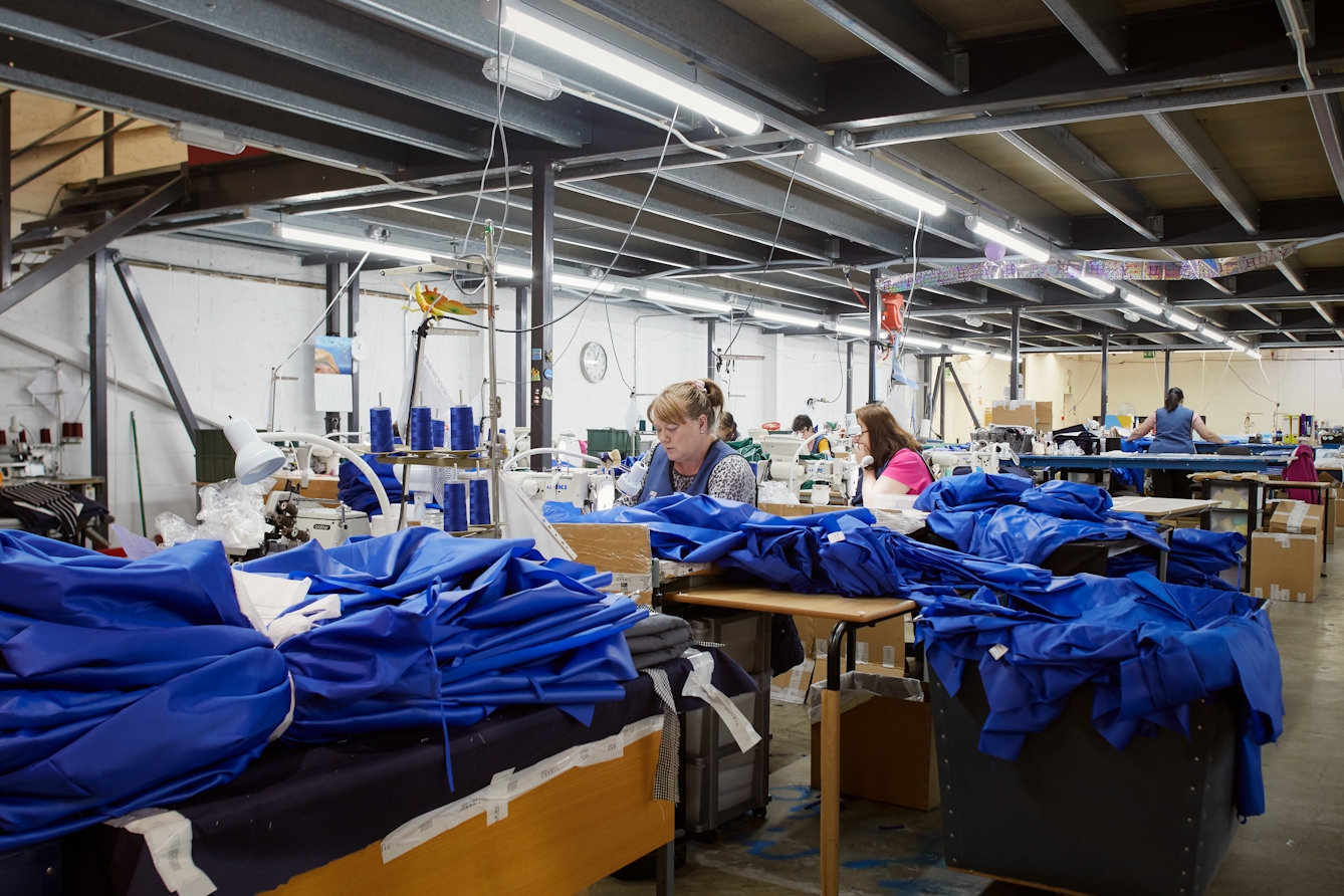 Photograph of a series of workstations within a clothing manufacturing workshop. There is a woman at the centre of the image working at a sewing machine. She is surrounded my blue medical fabric at various stages of construction.