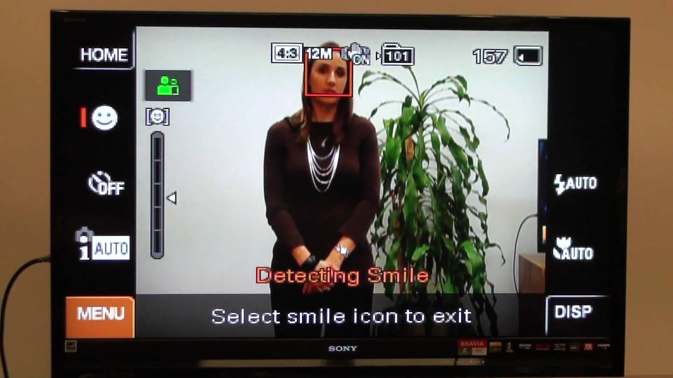 A screengrab from a YouTube video demonstrating the interface from a Sony Cybershot camera. On the camera screen a woman stands beside a plant and the camera screen reads "Detecting Smile".