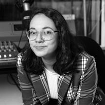 Photograph of a woman with dark, shoulder-length hair and transparent-rimmed glasses, leaning forward in a chair, with an audio mixing desk in the background.