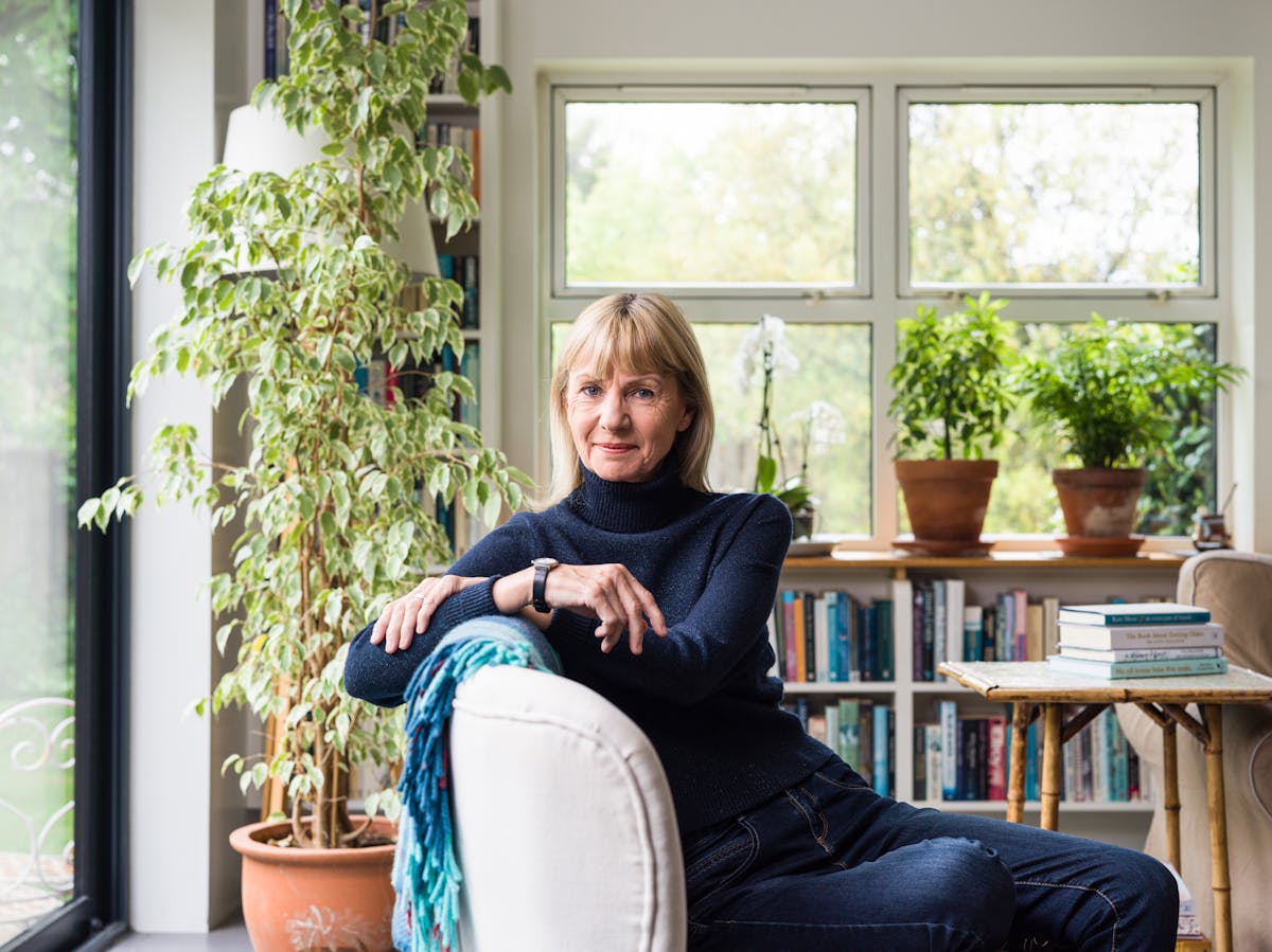 A photographic portrait of a woman with blonde hair wearing a navy turtleneck jumper and blue jeans. She is leaning to the left over the back of a sofa and in the background are a number of houseplants and shelves full of books.