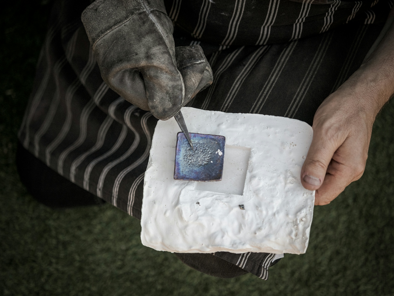 Photograph if a gloved right hand, pliers in hand holding the now solid piece of lead above the plaster-cast mould. The left hand is holding the plaster mould.