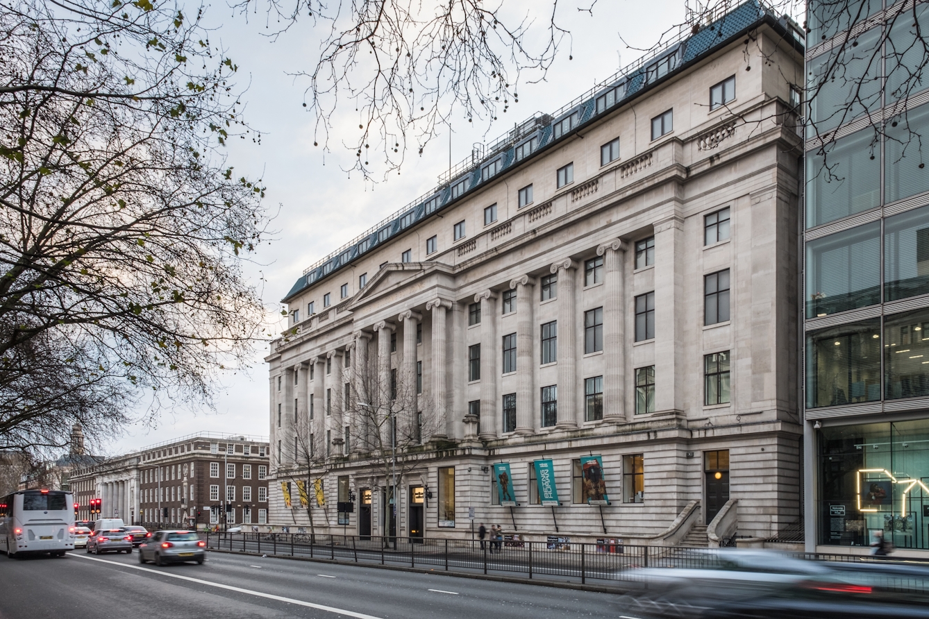 A photograph showing the front of the Wellcome Collection building, a grand building built in 1936, and Euston Road in front of it. The pale stone facade features 12 decorative columns, of which the four central columns support a triangular roof like decoration between the fourth and fifth floors. Six floors are visible, and each floor has fifteen windows, except the ground floor where there are three doors at street level.