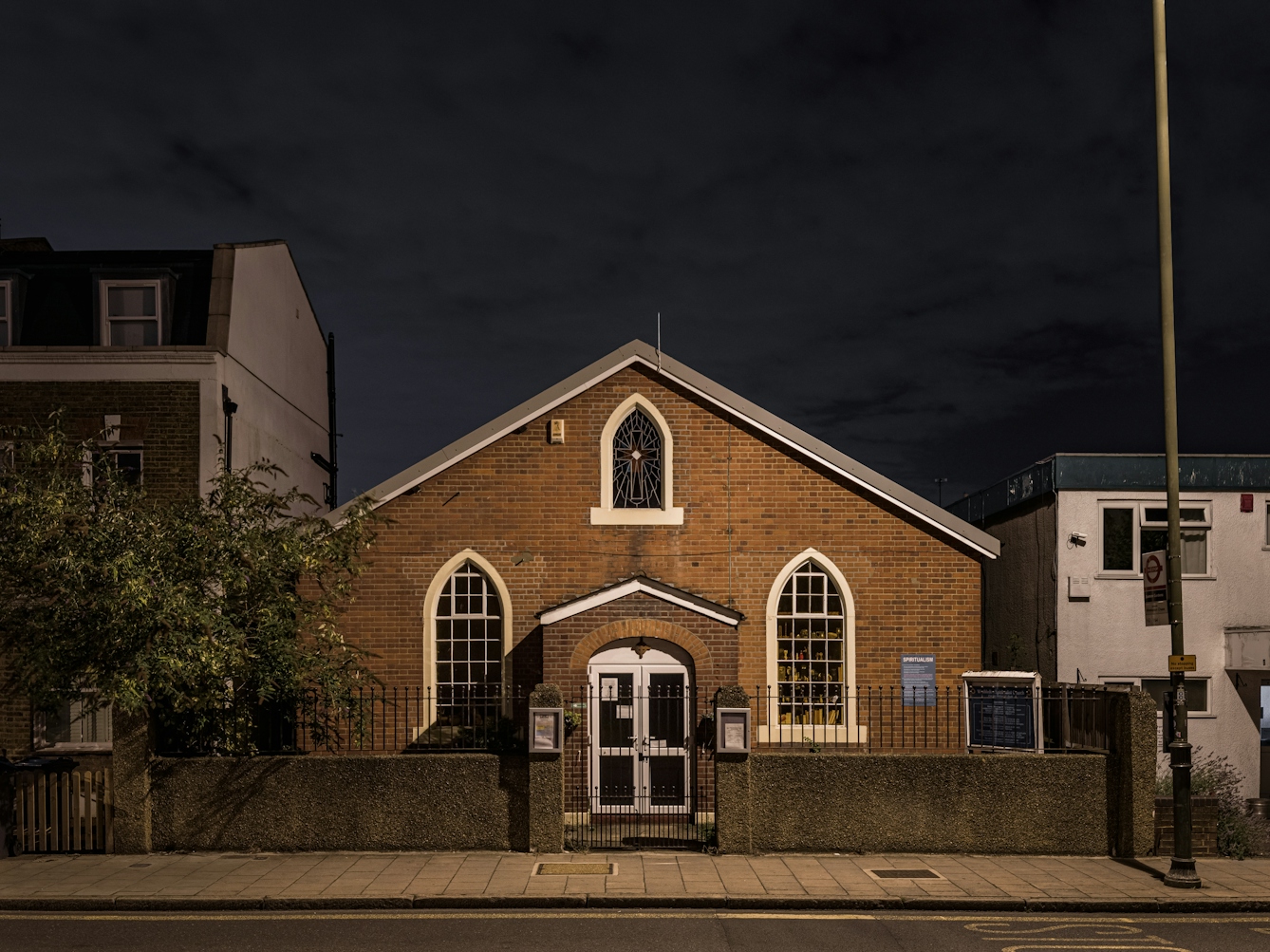 Photograph of Wimbledon Spiritualist Church at night.  The red brick Victorian building has white window recesses and a glass stained window bearing a cross in the apex of the facade.  The building is detached in a residential setting.