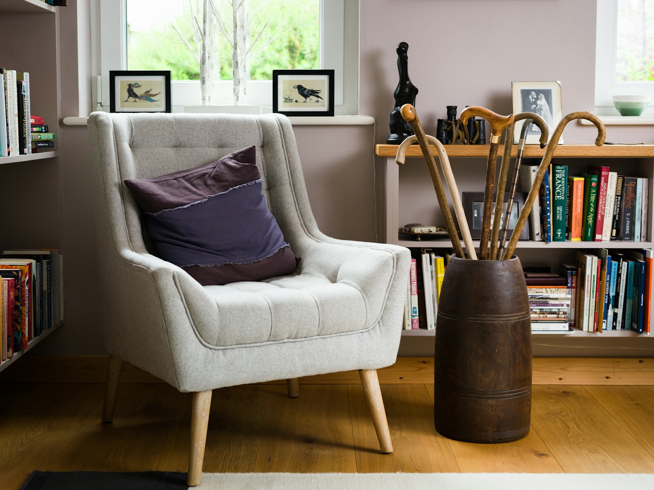 A photograph of a corner of a room with an armchair and round cylinder shaped wood container being used to hold a number of wooden walking sticks. In the background are shelves full of books and family photos in frames.