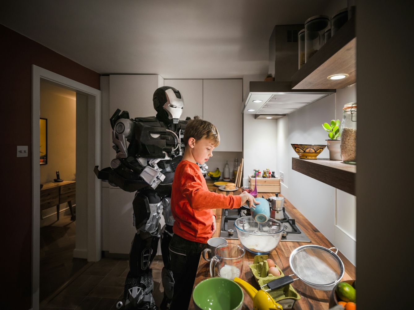 Photograph of a young boy standing at a kitchen countertop mixing the ingredients for pancakes. Behind him stands a large robot holding a spatular, ready to help make the pancakes.