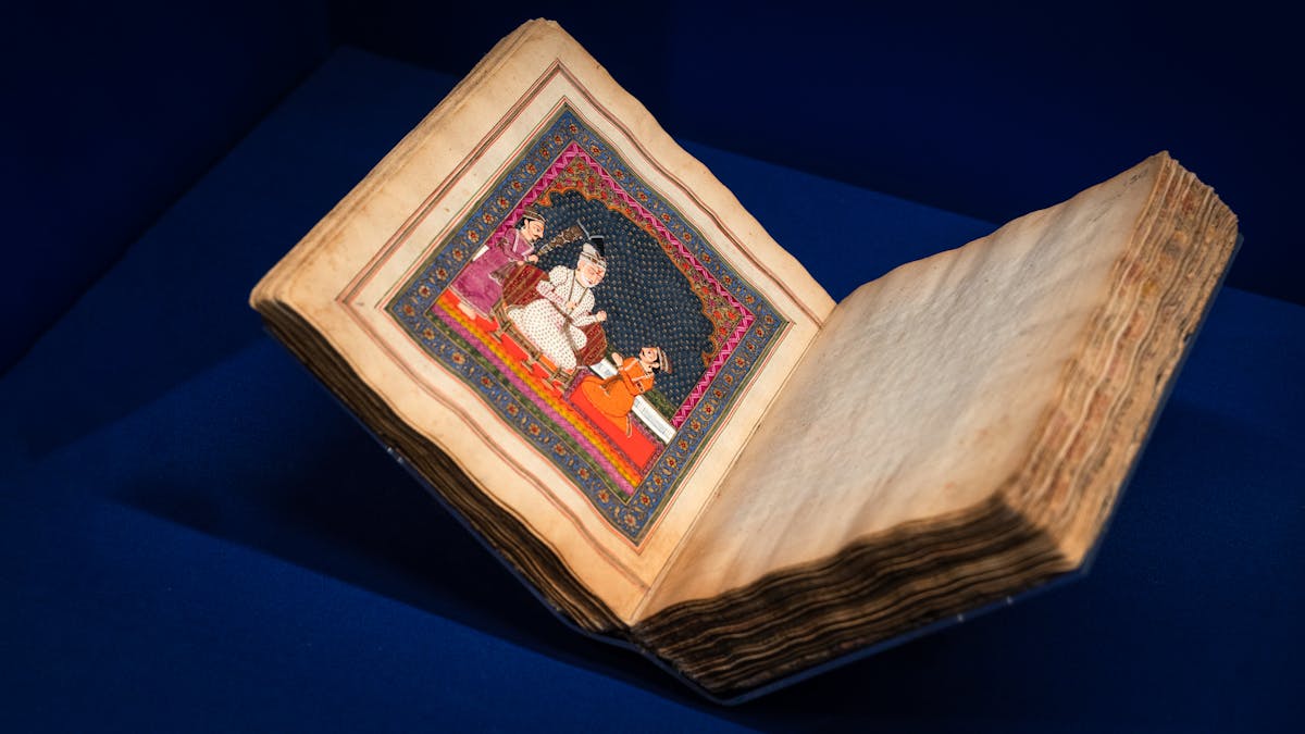 Photograph of an ornately illustrated Panjabi manuscript. The manuscript is pictured resting open, in an exhibition display case, with the illustration on the lefthand page and a blank page on the right. It is resting on a dark blue fabric background. The illustration is intricately detailed in vibrant colours, including reds, oranges and blues. It depicts a scene where a central character in white is talking with an individual in orange, kneeling in-front of him. The central character is being fanned by an individual in purple to the left.