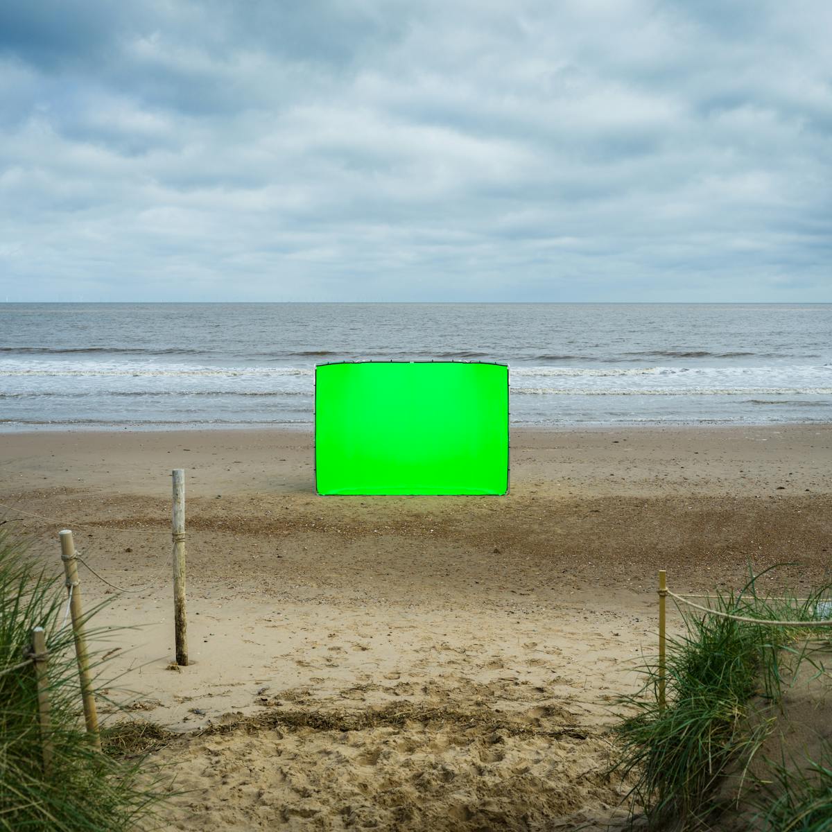 Colour landscape photograph showing a beach scene. The scene is split in half horizontally, with a blue sky and cloud covered upper and a sandy beach lower, with a strip of sea and breaking waves across the middle. In the centre of the image, in the middle distance is a large rectangular photographic background frame standing up vertically. Stretched across the frame is a vibrant green chroma key fabric. In the foreground is a path leading out to the beach from the sand dunes. The dunes rising up and covered in long green grass can be seen to the left and right of the scene. There are several wooden posts stuck in the ground with rope tied between them, protecting the dunes from access.