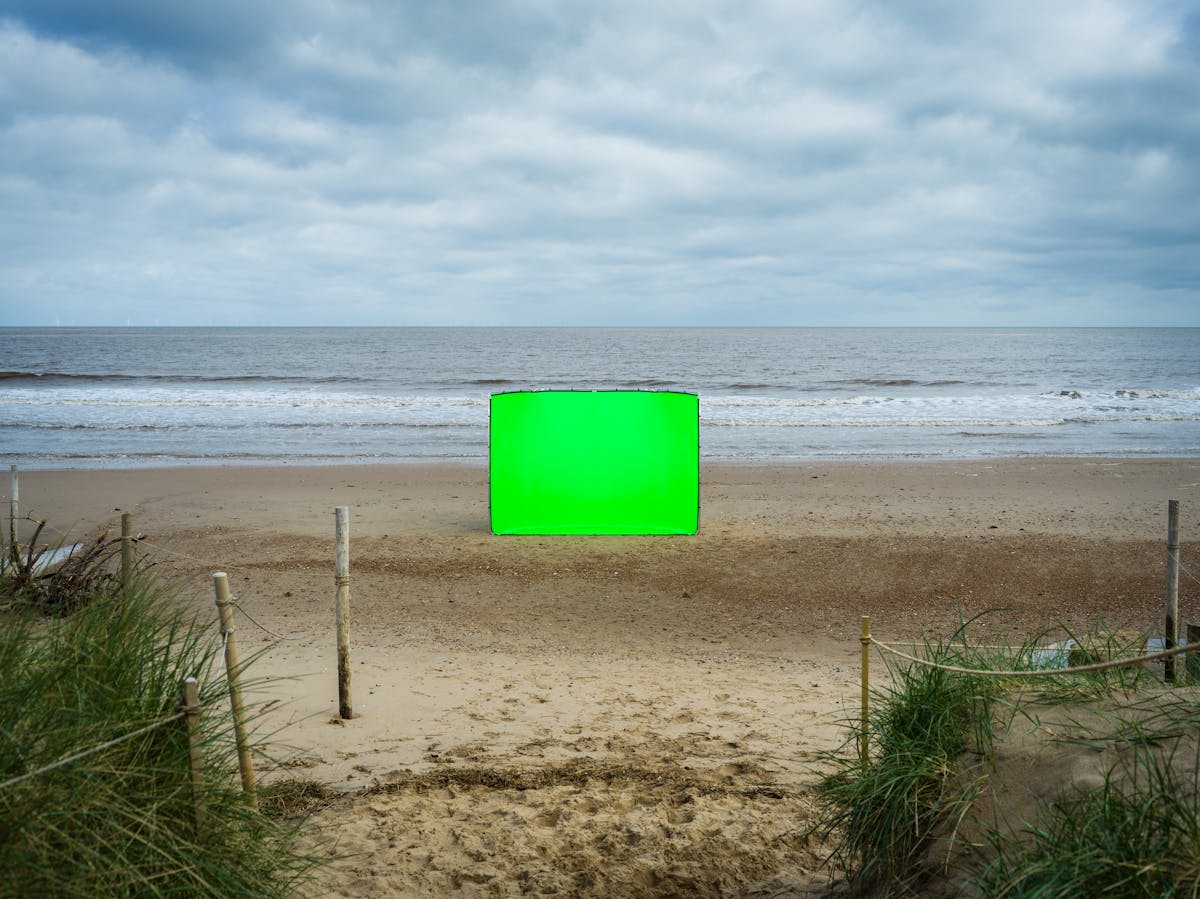 Colour landscape photograph showing a beach scene. The scene is split in half horizontally, with a blue sky and cloud covered upper and a sandy beach lower, with a strip of sea and breaking waves across the middle. In the centre of the image, in the middle distance is a large rectangular photographic background frame standing up vertically. Stretched across the frame is a vibrant green chroma key fabric. In the foreground is a path leading out to the beach from the sand dunes. The dunes rising up and covered in long green grass can be seen to the left and right of the scene. There are several wooden posts stuck in the ground with rope tied between them, protecting the dunes from access.