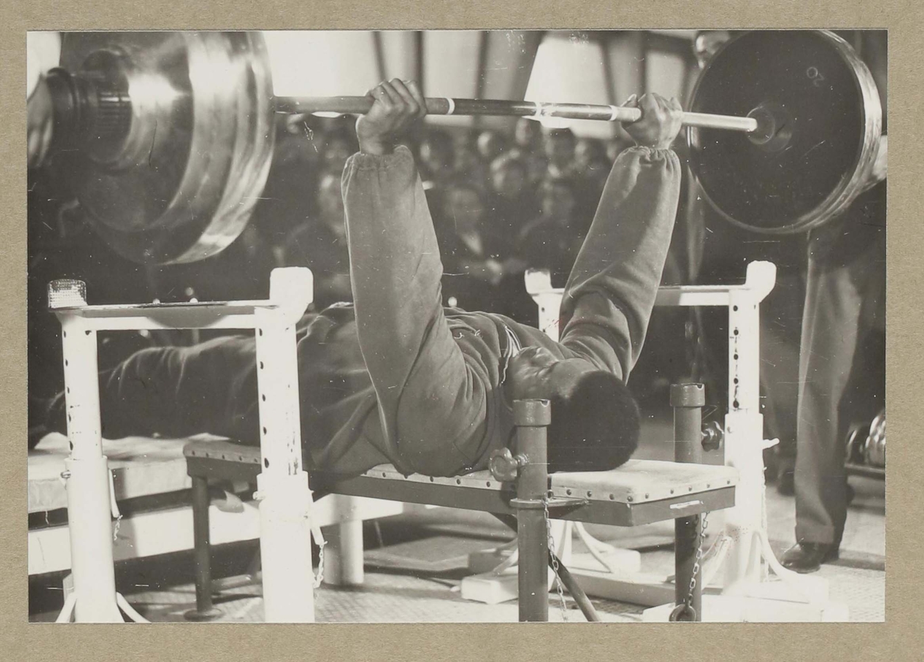 Black and white photographic print showing a black man on a weightlifting bench holding a large barbell above him with straight arms and clenched fists. A leg of a spotter or observer is just visible to the right, and behind the barbell and the weightlifter are an audience whose blurred faces lean forward, focusing on the competitor.