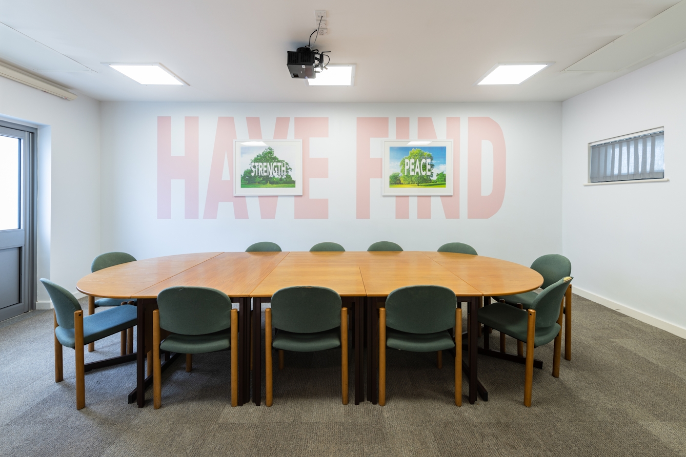 A photograph showing a white room containing a large table surrounded by chairs and a wall on which there is a large mural with the words ‘have’ and ‘find’ painted in large light pink capital letters. In the middle of the word ‘have’ is a smaller picture of a tree with the word ‘strength’ across it. In the middle of the word ‘find’ is a picture of a tree with the word ‘peace’ across it. 