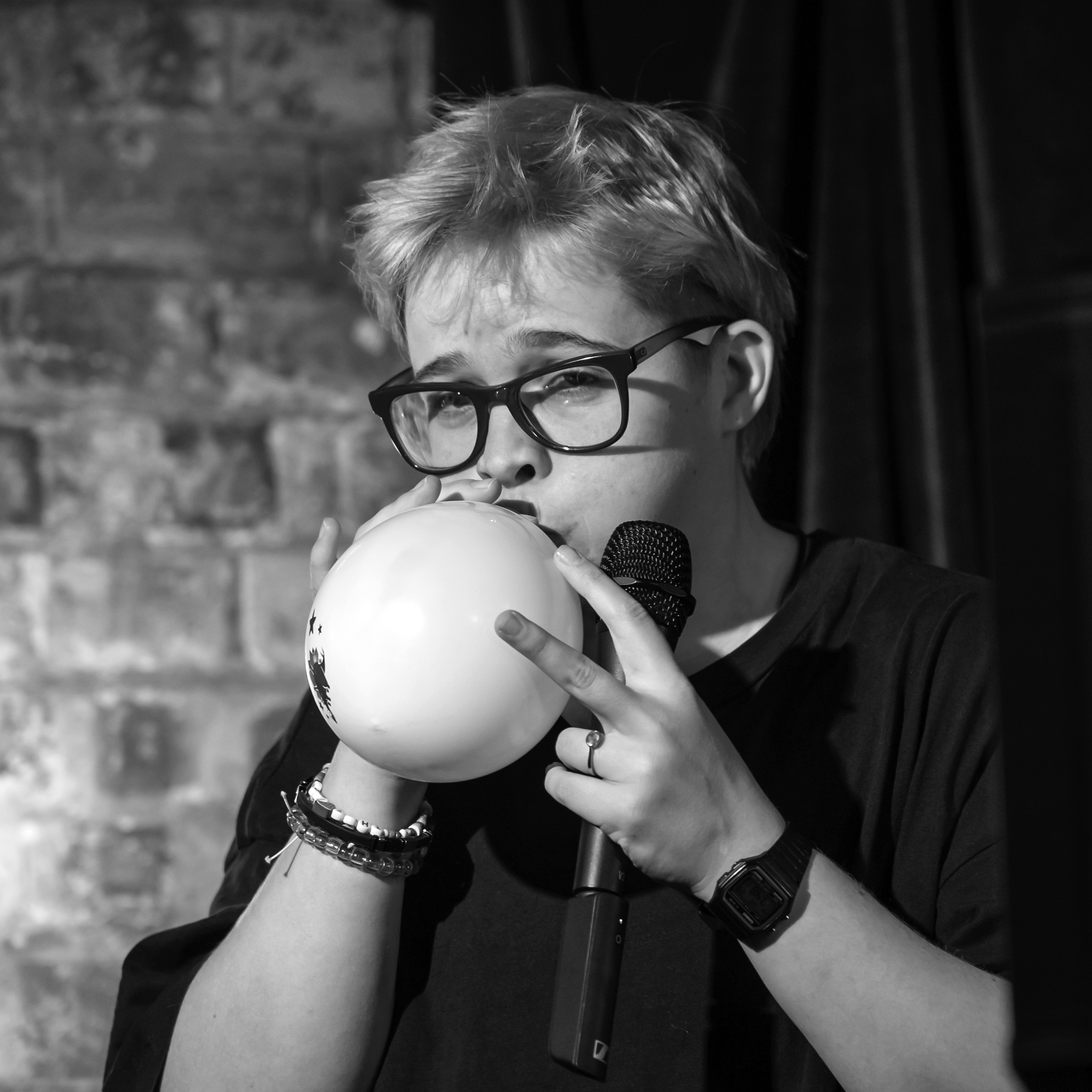 Photograph of a woman with short hair and glasses blowing up a balloon ad holding a stage microphone. 