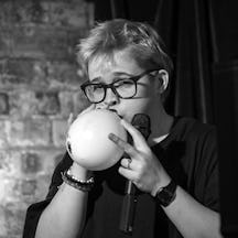 Photograph of a woman with short hair and glasses blowing up a balloon ad holding a stage microphone. 