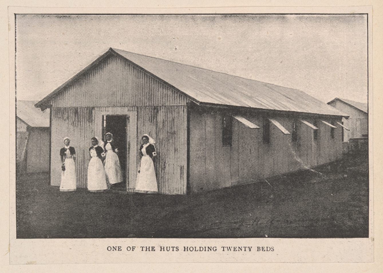 Black and white photo of a metal hut with four nurses standing outside the front. It is captioned: 'One of the huts holding twenty beds'.