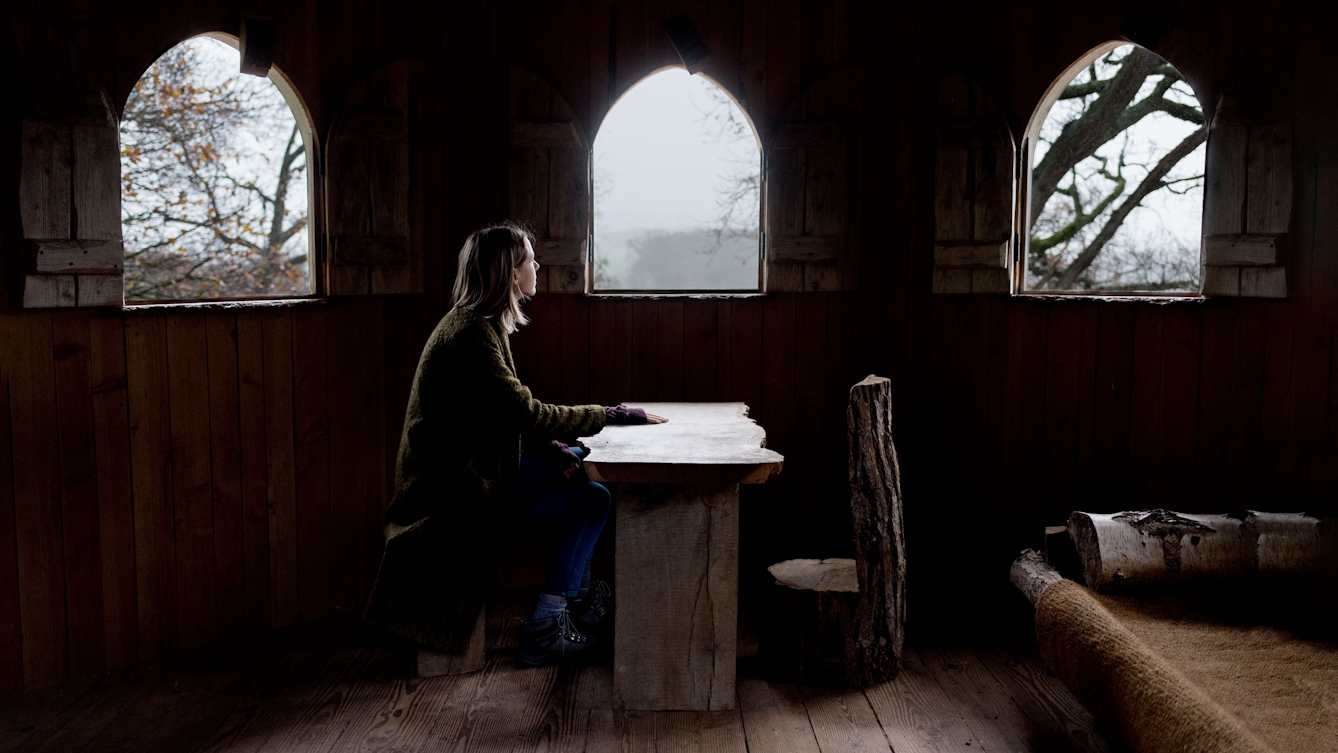 Photograph of a young woman sitting at a wooden table in a hermitage. On the other side the table is an empty chair carved from a tree trunk. behind her are three small arched windows which show the trees and mist outside.