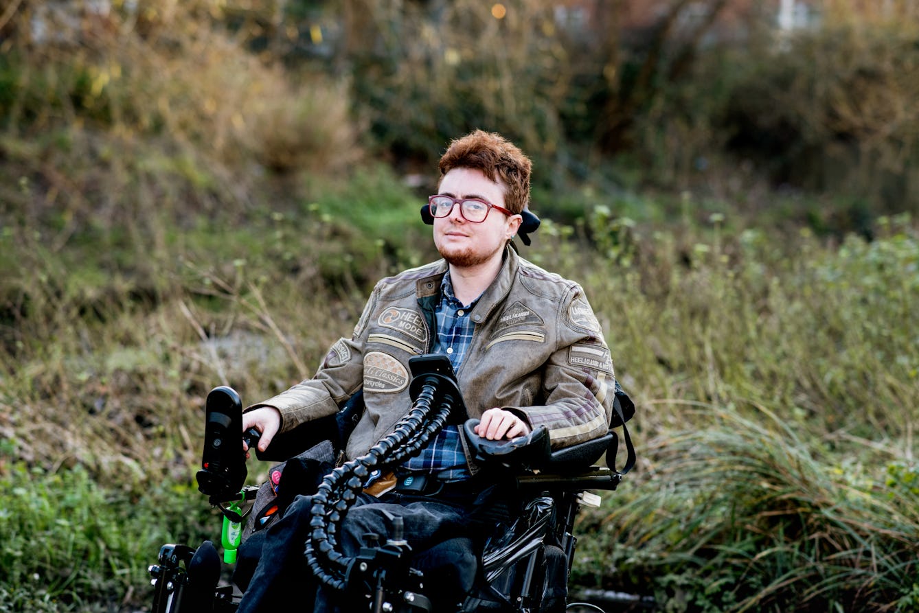 Photographic portrait of a young person seated outside in a electric wheelchair looking to camera. Behind them are long grasses and shrubs lit from behind by a low sun.

