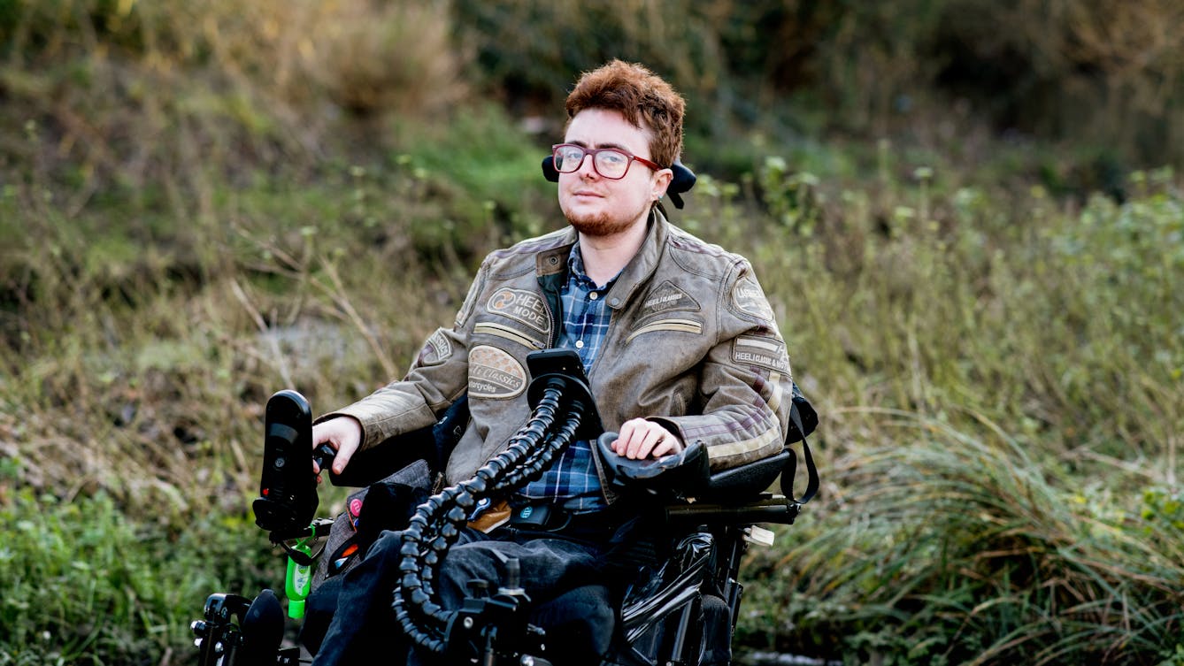 Photographic portrait of a young person seated outside in a electric wheelchair looking to camera. Behind them are long grasses and shrubs lit from behind by a low sun.
