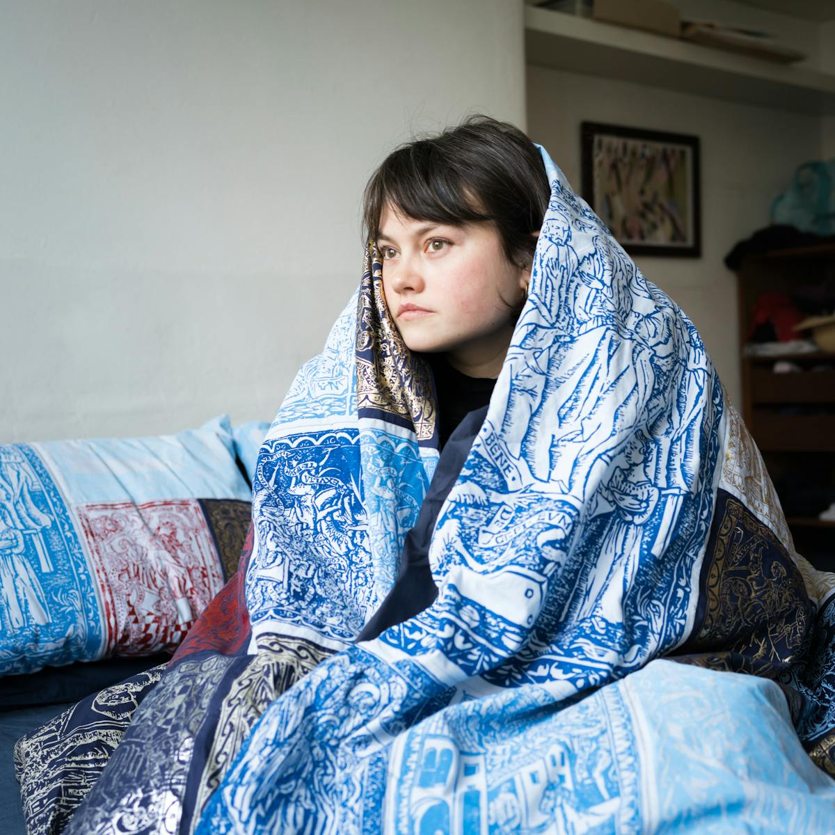 Photograph of a young woman sitting on her bed, wrapped in her duvet. The duvet cover is made of a patchwork quilt of blue, red and gold screen prints depicting woodblock engravings from a manuscript. Behind her is a framed photograph on the wall, shelves and part of her wardrobe.