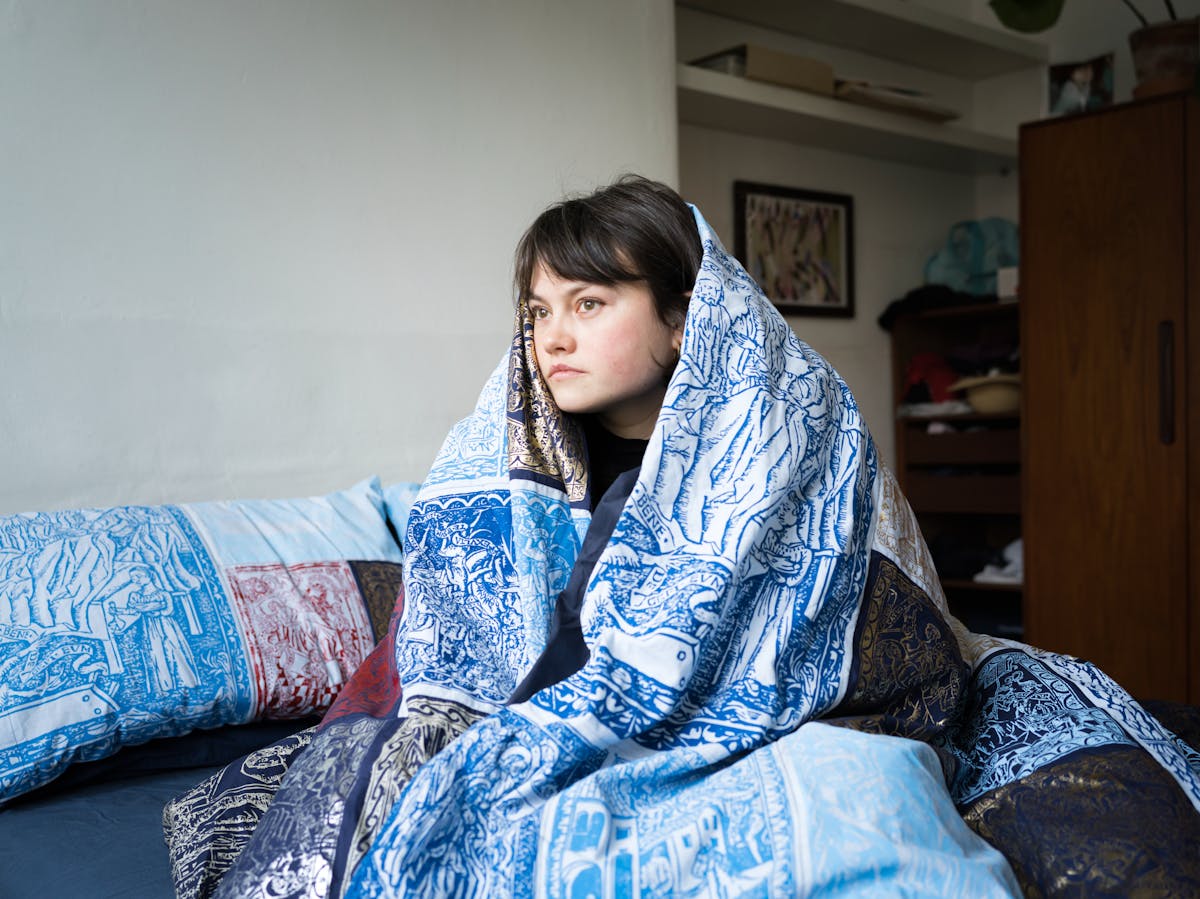 Photograph of a young woman sitting on her bed, wrapped in her duvet. The duvet cover is made of a patchwork quilt of blue, red and gold screen prints depicting woodblock engravings from a manuscript. Behind her is a framed photograph on the wall, shelves and part of her wardrobe.