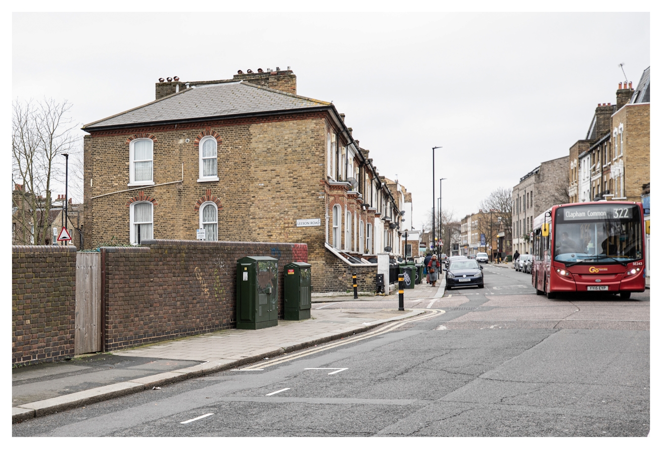 Photograph of a South London street scene in Brixton, showing residential buildings on either side. On the road is a single decker red London bus with the number 322 and destination 'Clapham Common' written on the front.