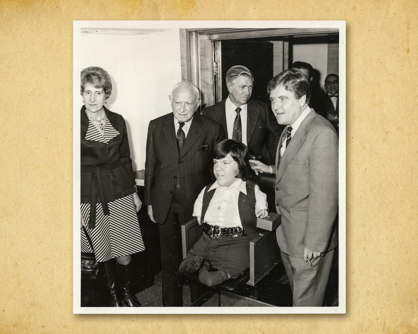 Photograph of a black and white photographic print, resting on a brown paper textured background. The print shows a young man seated in the centre of the image who has short arms and legs as a result of his mother being prescribed thalidomide during pregnancy. The young man is surrounded by a woman and three older men in suits and ties.