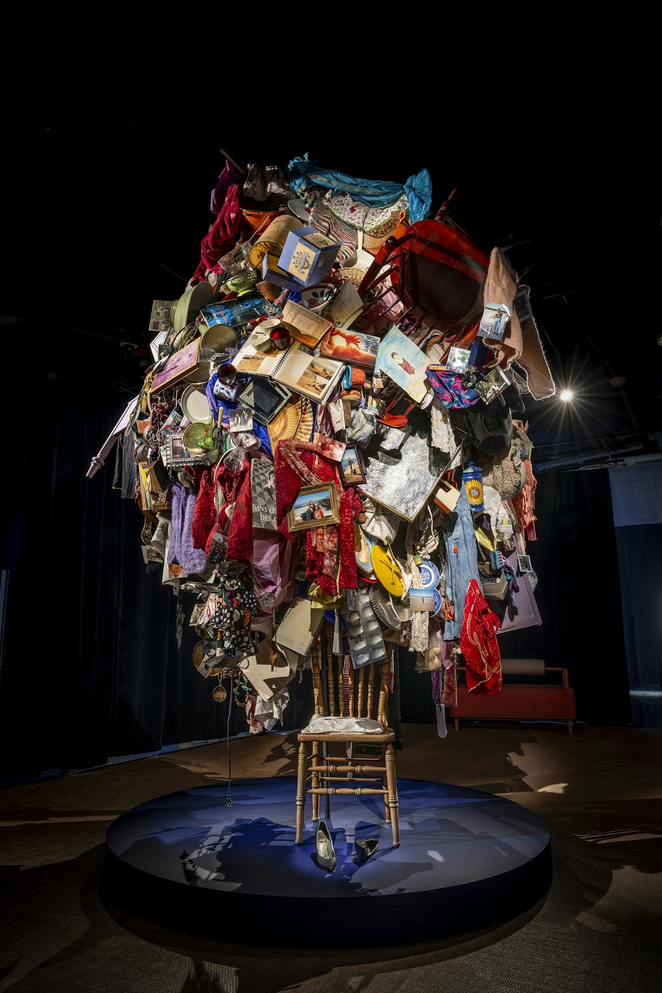 An artwork on display on a low circular plinth. The work consists of a dining chair with a mass of clothes, books, pictures and other domestic objects piled pricariously on top of the chair.