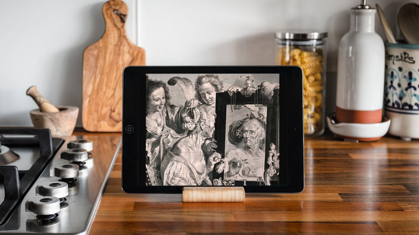 An iPad on a stand in a kitchen. On the screen is an etching of an old lady sitting at her dressing table mirror holding flowers, two younger women are putting feathers in her hair.