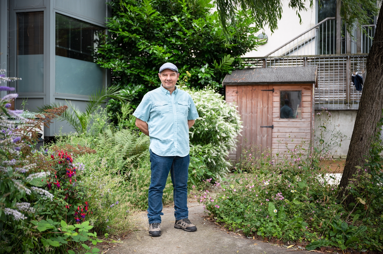 Photograph of a man in full length standing outside in a garden like environment. He is wearing a flat cap, light blue short sleeved shirt and a pair of blue jeans. His hand are clasped behind his back and he is looking straight to camera. Either side of the path he is standing on are green shrubs in bloom with colourful flowers. In the distance is a small wooden shed and a glass and grey metal building.