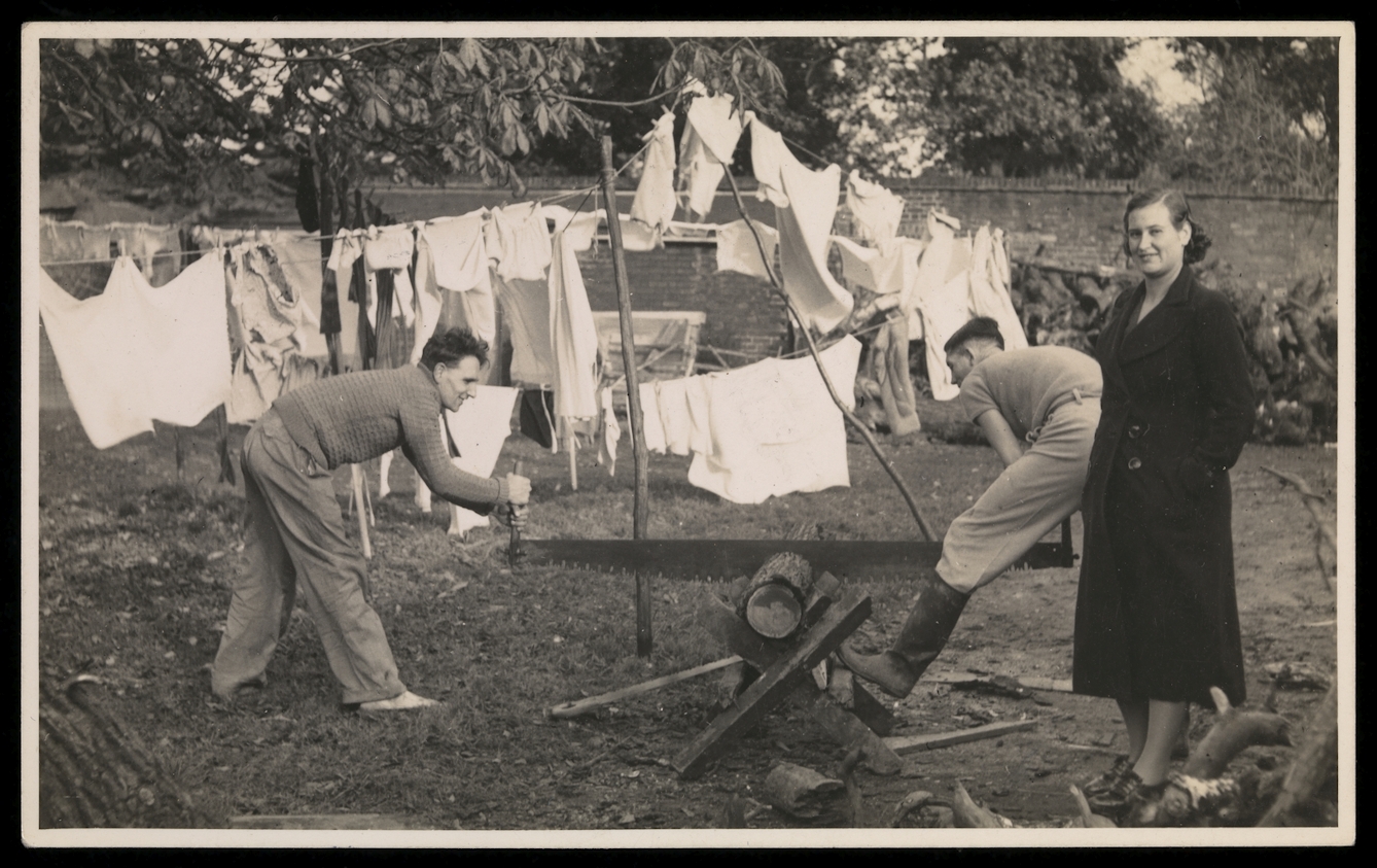 Black and white photograph showing tow men sawing a log in half, with a women in the foregound facing the camera, and washing lines in the background.