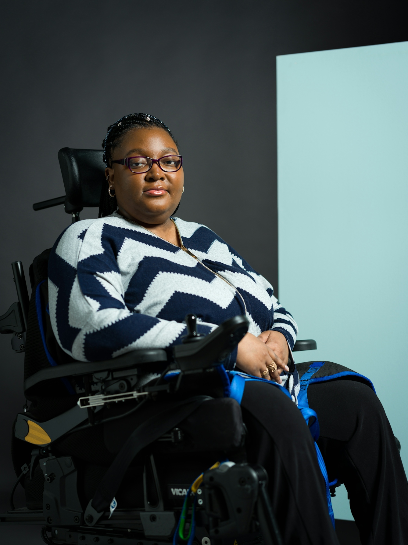 Colour photograph three-quarter length portrait of Matilda Feyiṣayọ Ibini, who is a black woman in a motorised wheelchair. She has braided hair, rectangular black glasses, a zig-zag navy and white jumper, and black trousers. She wears gold earrings and gold rings on her fingers.