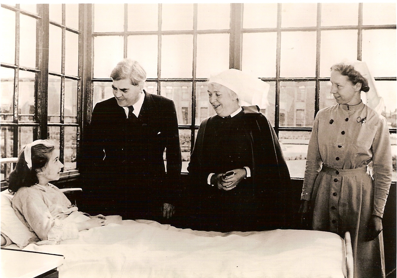Black and white photograph showing a girl sitting up in a hospital bed. She is looking up and smiling. To the girl's left are three people in a line next to the bed. Closest to the girl is a man in a suit looking down at the girl and smiling. To his left is a woman wearing a black cloak and white headdress. She is smiling and looking down at the girl. To her left there is another woman wearing a button up dress and white headdress. She is also smiling and looking towards the girl.