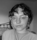 Black and white photograph of the head and shoulders of a young woman with short dark hair and large round glasses. She is looking directly at the viewer.